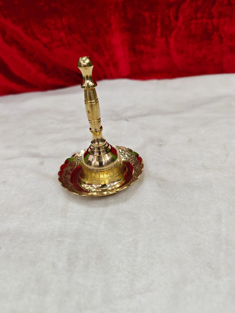 May include: A polished brass bell featuring a slender, multi-ringed handle and a wide, circular base with an ornate, scalloped edge. The metal surface exhibits a bright, reflective gold tone, showcasing intricate engravings along the rim.