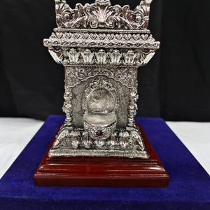 Puede incluir: Un objeto decorativo de color plateado que presenta una estructura arquitectónica ornamentada con intrincados patrones en relieve, centrado por un pequeño cuenco, todo montado sobre una base de madera pulida de color marrón rojizo.