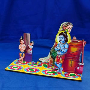 Puede incluir: Un expositor decorativo de mesa con una deidad de piel azul atada a un gran mortero de madera, acompañada de una figura femenina y dos figuras más pequeñas junto a un pilar, sobre una base con un patrón floral en amarillo y rojo.