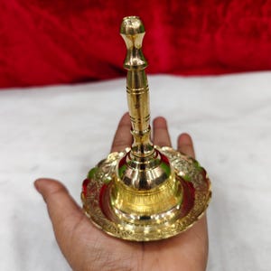 May include: A polished brass bell featuring a tall, slender handle with horizontal grooves and a wide, circular base. The base is decorated with an intricate, embossed floral pattern along its scalloped edge, creating a reflective metallic finish.