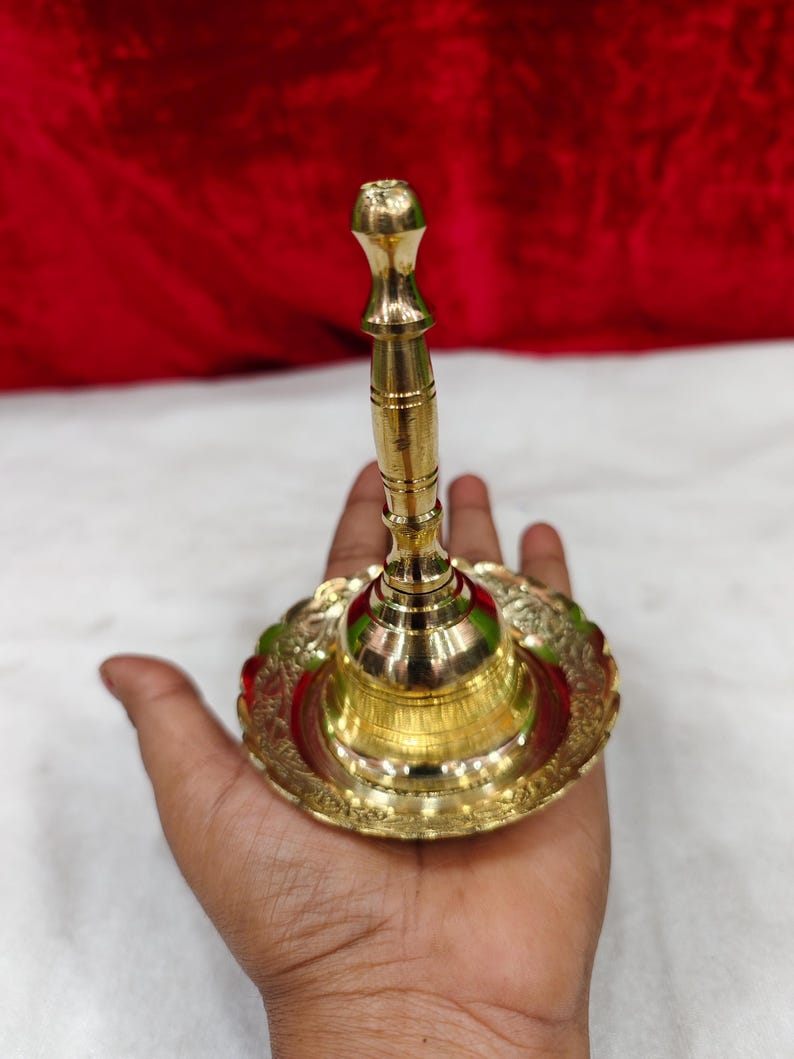 May include: A polished brass bell featuring a tall, slender handle with horizontal grooves and a wide, circular base. The base is decorated with an intricate, embossed floral pattern along its scalloped edge, creating a reflective metallic finish.