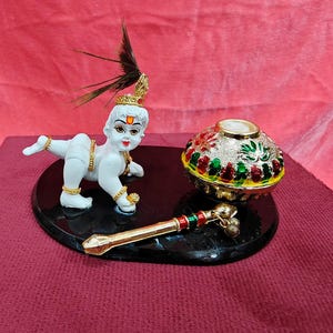 May include: A white figurine of a crawling child with a peacock feather headpiece, a gold-toned flute with small bells, and a decorative metallic bowl with red and green accents, all mounted on a smooth, oval-shaped black base.