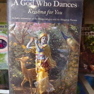 Puede incluir: Un libro titulado A God Who Dances Krishna for You de Carl Woodham. La portada muestra a una figura de piel azul tocando la flauta, vistiendo prendas amarillas y una guirnalda floral, junto a un ciervo y un pavo real en un bosque.