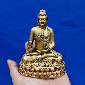Könnte beinhalten: Eine goldfarbene Buddha-Statue in sitzender Lotushaltung mit detaillierten Gewandverzierungen und einer erhobenen rechten Hand als Schutzgeste. Die Figur ruht auf einem dekorativen Sockel, der rundum mit geschnitzten Lotusblütenmustern verziert ist.