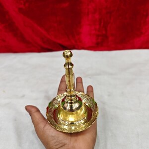 May include: A polished brass hand bell featuring a slender, tiered handle and a wide, circular base with an ornate, scalloped edge. The metal surface is reflective and smooth, showcasing a metallic gold tone throughout the entire object.