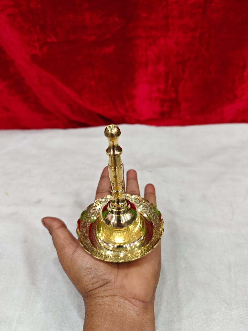 May include: A polished brass hand bell featuring a slender, tiered handle and a wide, circular base with an ornate, scalloped edge. The metal surface is reflective and smooth, showcasing a metallic gold tone throughout the entire object.