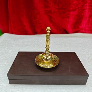 May include: A polished brass hand bell with a slender, tiered handle and a wide, flared base featuring intricate engraved patterns. The bell is positioned upright on a rectangular, dark brown box with a smooth, matte finish and clean edges.