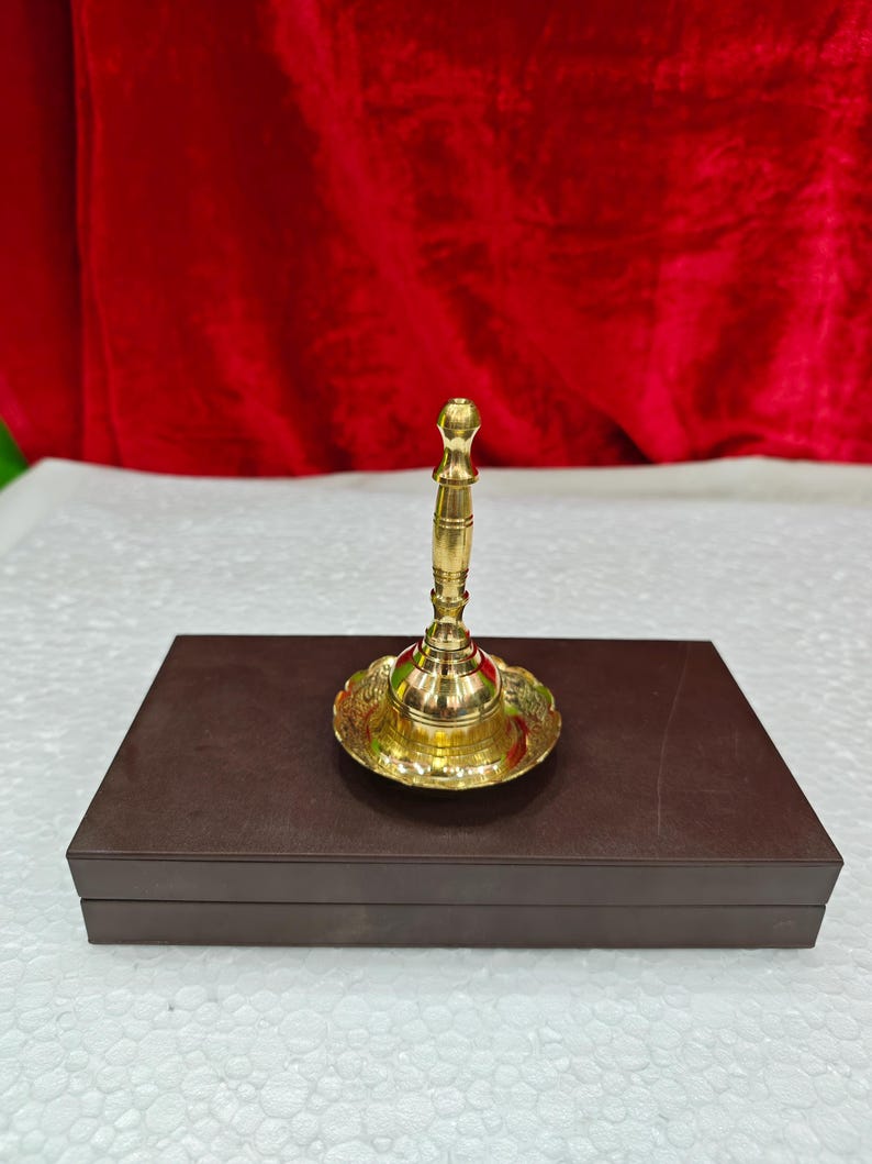 May include: A polished brass hand bell with a slender, tiered handle and a wide, flared base featuring intricate engraved patterns. The bell is positioned upright on a rectangular, dark brown box with a smooth, matte finish and clean edges.
