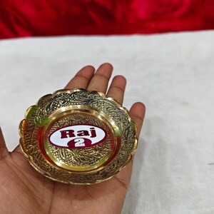 May include: A small, round, gold-colored brass dish with a scalloped edge and intricate embossed floral patterns. The center of the dish features a white oval sticker with the red text Raj 2 printed in a bold, sans-serif font.