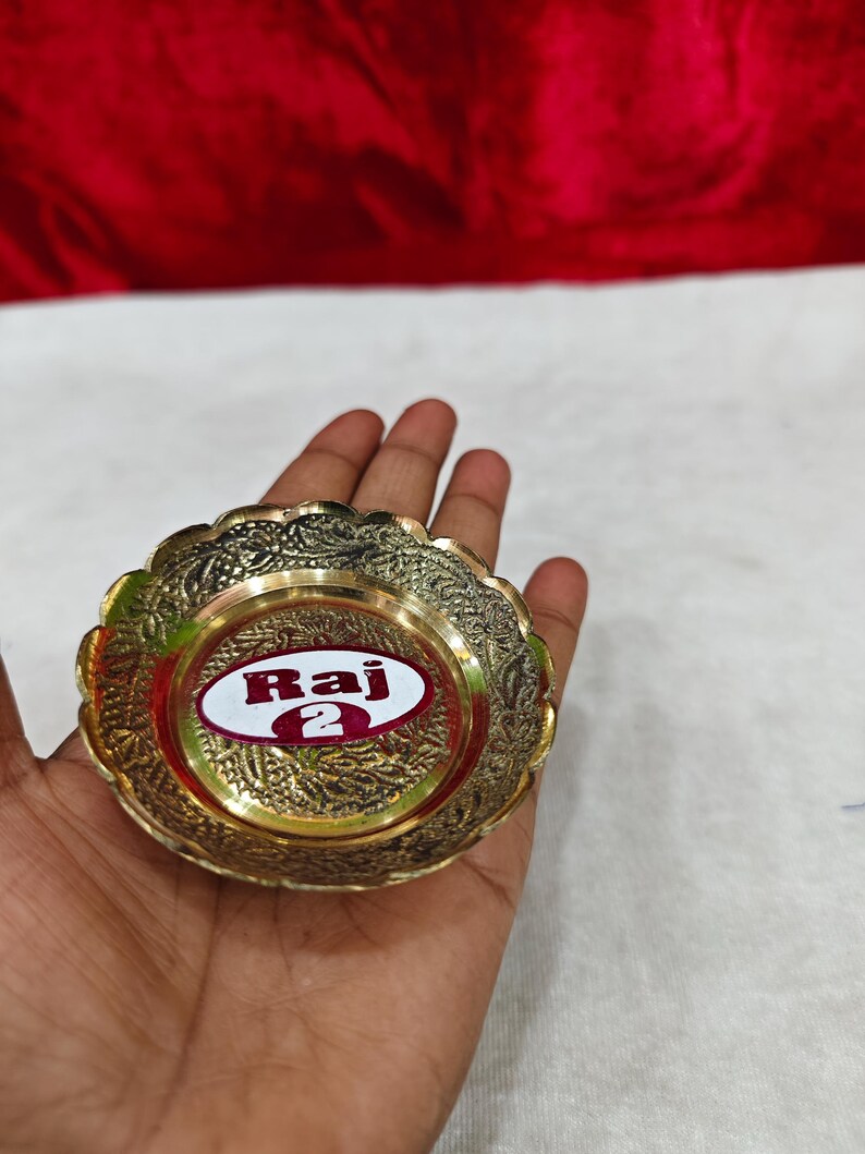 May include: A small, round, gold-colored brass dish with a scalloped edge and intricate embossed floral patterns. The center of the dish features a white oval sticker with the red text Raj 2 printed in a bold, sans-serif font.