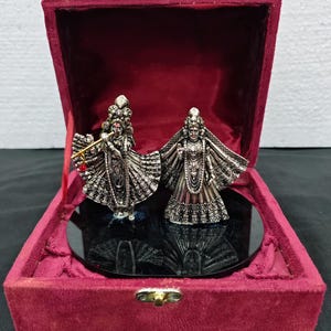 Puede incluir: Dos figuras de metal en tono plateado de deidades, una tocando una flauta de color dorado y la otra de pie al lado, ambas con detalles intrincados en la vestimenta, colocadas sobre una base circular negra dentro de una caja forrada de terciopelo rojo.