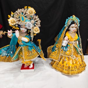 Puede incluir: Dos figuras ornamentadas de deidades vestidas con telas vibrantes en amarillo y turquesa con intrincados bordados dorados, lentejuelas y abalorios. La figura de la izquierda sostiene una flauta dorada, mientras que la derecha lleva un tocado y velo.
