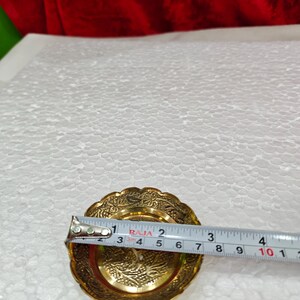 May include: A small, round, gold-colored metal dish with an intricate embossed floral pattern along the scalloped rim and center. A measuring tape is placed across the dish, showing a diameter of approximately 3.5 inches at its widest point.