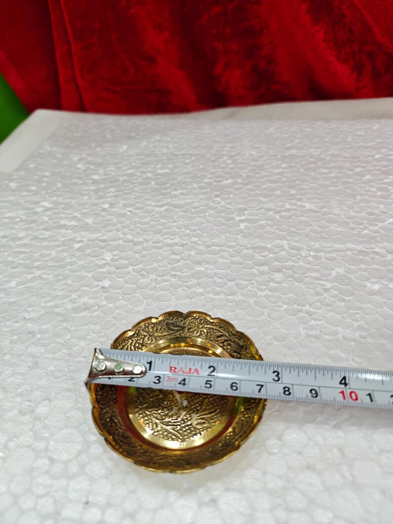 May include: A small, round, gold-colored metal dish with an intricate embossed floral pattern along the scalloped rim and center. A measuring tape is placed across the dish, showing a diameter of approximately 3.5 inches at its widest point.