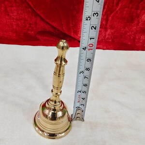 May include: A polished brass hand bell with a slender, tiered handle and a flared base. A metal measuring tape is positioned vertically next to the bell, indicating a total height of approximately 5.5 inches from the base to the top handle.