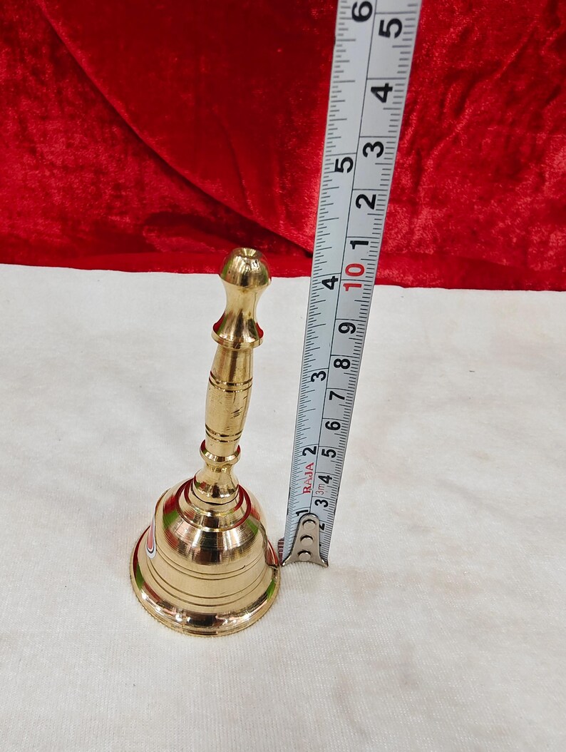 May include: A polished brass hand bell with a slender, tiered handle and a flared base. A metal measuring tape is positioned vertically next to the bell, indicating a total height of approximately 5.5 inches from the base to the top handle.