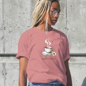 Könnte beinhalten: Rosafarbenes T-Shirt mit einer Grafik einer dampfenden Kaffeetasse auf einem Buch, einer Blume und dem Schriftzug "soft reset". Das T-Shirt ist einfarbig und hat hochgekrempelte Ärmel. Die Person trägt Jeans.