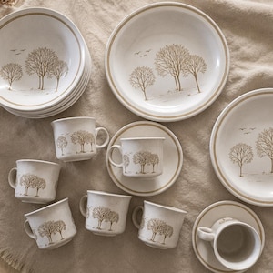 Puede incluir: Un juego de vajilla de cerámica color hueso con platos, cuencos y tazas decorados con un patrón de líneas marrones de árboles y pájaros. El conjunto incluye una pila de cuencos, tres platos grandes y cinco tazas, algunas con platillos.