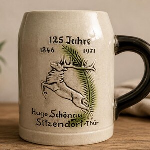 Może przedstawiać: Beżowy ceramiczny kufel do piwa z ciemnobrązowym uchem. Z przodu znajduje się wypukły wizerunek skaczącego jelenia, zielony liść palmy oraz napis 125 Jahre 1846 1971 Hugo Schönau Sitzendorf-Thür. Kufel ma gładkie wykończenie i cylindryczny kształt.