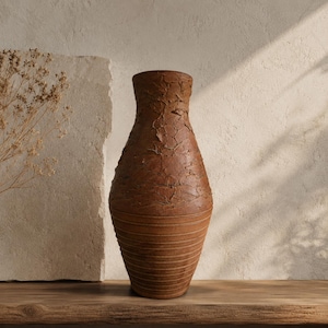 Può includere: Un alto vaso in ceramica marrone con una superficie ruvida e screpolata nella parte superiore e linee orizzontali scanalate attorno alla base. Il vaso presenta un collo stretto che si allarga in un corpo arrotondato, finitura rustica e terrosa.