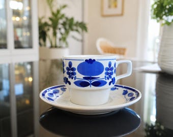 Könnte beinhalten: Ein weißes Tassen- und Untertassen-Set aus Keramik mit einem sich wiederholenden blauen Blumen- und Pflanzenmuster. Die Tasse hat einen zylindrischen Körper mit abgestuftem Boden und einem Henkel, während die Untertasse ein passendes blaues Motiv zeigt.