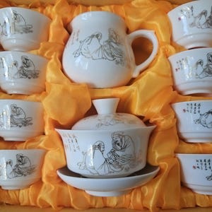 May include: A white ceramic tea set featuring a teapot, a lidded bowl with a saucer, and seven small cups. Each piece is decorated with black line drawings of figures and Chinese calligraphy characters, all arranged within a bright yellow fabric lining.