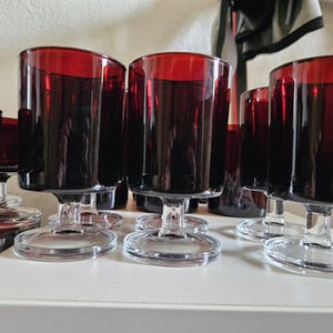 May include: A collection of deep red glass goblets with clear glass stems and circular bases. Each goblet features a cylindrical bowl with a smooth, translucent finish, arranged in a group to showcase the consistent color and structural design.