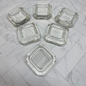 May include: A set of six clear glass square coasters with beveled edges and a hollow center. Each coaster features a thick, transparent frame with a smooth, polished finish, arranged in a cluster to display their uniform shape and design.