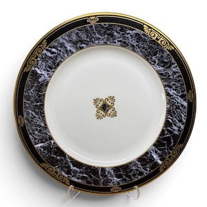 May include: A round porcelain dinner plate featuring a white center with a gold filigree emblem. The wide rim displays a black and white marble pattern bordered by a gold edge and accented with repeating gold geometric and floral motifs.