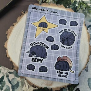 May include: A rectangular sticker sheet with a light blue plaid background featuring various dark blue isopod illustrations. Text includes "silly pods n' puns", "me did it", "THEY SEE ME ROLLIN' THEY HATIN'", "ISO-FUCKIN EEPY", and "LEAF ME ALONE" in black lettering.