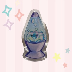 May include: A clear, acrylic charm in the shape of a lava lamp. The lamp features a blue and purple color scheme with a cartoonish creature inside. The background is a pink and white striped pattern with star and diamond shapes.