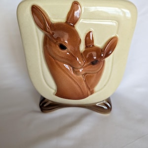 May include: A cream-colored ceramic vase featuring a raised, glossy brown relief of a deer and a fawn. The vase has a wide, flat top and a curved base, with the deer and fawn depicted in a side-profile embrace against a smooth, light background.