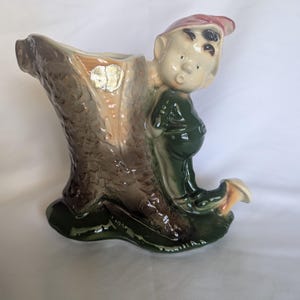 May include: A ceramic planter shaped like a hollow tree stump with a textured brown bark finish. A small figure wearing a green outfit and a pink cap leans against the side of the stump. The base is dark green and features a small mushroom detail.