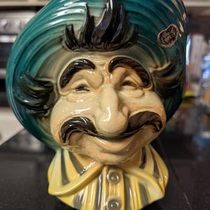 May include: Vintage ceramic figurine of a smiling man with a large mustache and a turquoise hat. The figurine has a yellow and black striped shirt and the words "Royal Copley" on a sticker.