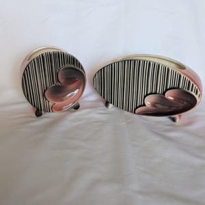 May include: Two ceramic vases with a rounded and oval shape, featuring a textured surface with vertical black lines and smooth, glossy pink accents. Each vase is supported by small, dark feet, showcasing a distinct contrast between the matte-textured vertical stripes and the polished, curved pink elements on the front.