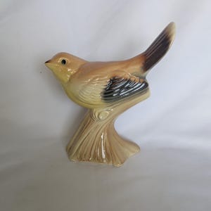 May include: A ceramic bird figurine perched on a textured tree stump base. The bird features a glossy finish with light brown, tan, and cream plumage, accented by dark brown wing and tail feathers, and a small black eye detail on its head.