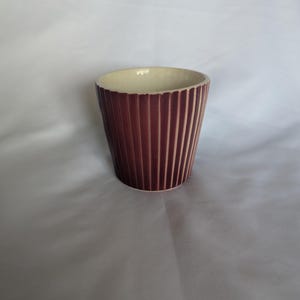 May include: A cylindrical ceramic planter featuring a deep burgundy exterior with vertical ribbed detailing. The interior of the vessel is finished in a smooth, off-white glaze, contrasting with the dark, textured outer surface of the pot.
