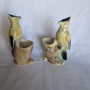 May include: Two ceramic bird-shaped planters featuring blue and yellow birds perched on brown tree stumps. The birds have dark blue wings and tails, while the tree stump bases are textured with molded floral details and a glossy finish.