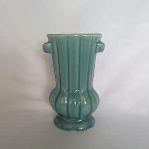 May include: A sage green ceramic vase featuring a vertical ribbed texture throughout the body. The vessel has a flared rim, a rounded base, and two small, protruding handles on opposite sides near the top, finished with a glossy glaze.