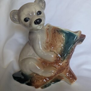 May include: A ceramic figurine of a light gray bear sitting against a textured brown and dark green tree stump. The bear has black eyes and a black nose, with a glossy finish. The tree stump features a hollow opening at the top, serving as a vase.