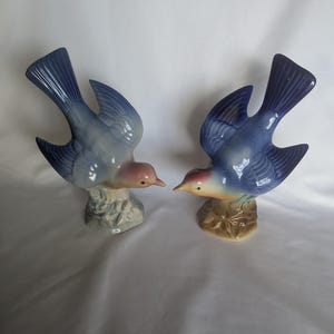May include: Two ceramic bird figurines with blue wings and tails, pinkish-tan heads, and dark eyes. Each bird is perched on a textured base. The left bird has a light blue base, while the right bird has a brown base. Both birds are facing each other.