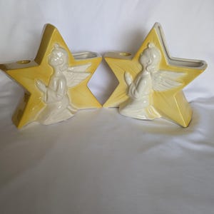 May include: Two ceramic candle holders shaped like five-pointed stars with a yellow gradient finish. Each star features a raised, white, kneeling angel figure with hands pressed together in prayer and wings extended, set against the star background.