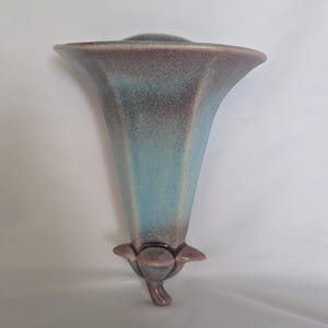 May include: A wall-mounted ceramic vase featuring a flared, trumpet-shaped opening. The piece displays a gradient glaze transitioning from a soft, dusty purple at the rim and base to a vibrant, light blue hue across the central body section.