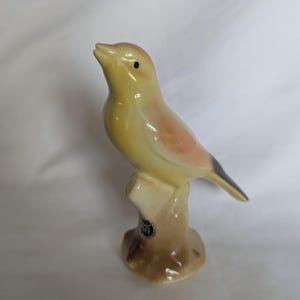 May include: A ceramic bird figurine perched on a brown tree stump base. The bird features a pale yellow body with soft orange-toned shading on the chest and wings, a small black eye, and a dark-tipped tail. A small circular sticker is visible on the base.