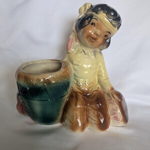 May include: A ceramic planter shaped like a figure kneeling beside a drum. The figure has dark hair, a light-colored shirt, and brown pants. The drum is dark green with a brown rim. The piece features a glossy finish and a hollow opening for plants.