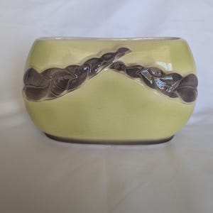 May include: An oval-shaped ceramic planter with a glossy light chartreuse green glaze. The exterior features a raised, dark brown relief pattern of intertwined leaves or vines that wrap across the front center of the vessel.