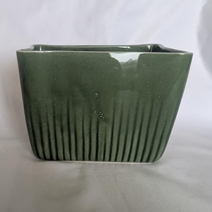May include: A rectangular ceramic planter with a glossy sage green glaze. The exterior features a series of vertical, recessed fluted grooves that run from the base toward the top rim, creating a textured appearance on the smooth, reflective surface.