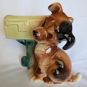 May include: A ceramic figurine featuring a brown and black dog sitting beside a pale yellow mailbox. The dog has a glossy finish, pointed ears, and a cream-colored chest. The mailbox is mounted on a teal post, creating a decorative piece.