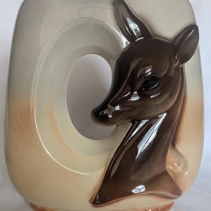 May include: A ceramic vase featuring a glossy, dark brown sculpted deer head relief on a cream and tan ombre background. The vase has a large, oval-shaped cutout opening on the left side and a decorative beaded texture along the bottom base edge.