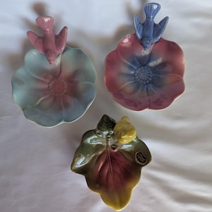 May include: Three ceramic trinket dishes. Two are flower-shaped bowls with birds perched on the rims: one in light blue and pink with a pink bird, and one in pink and blue with a blue bird. The third is a leaf-shaped dish in green and yellow with a yellow bird.